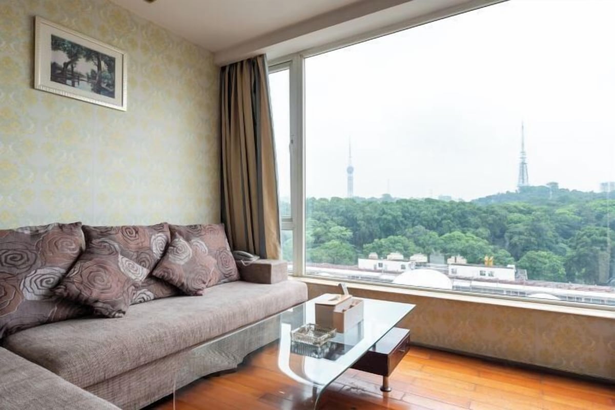A living room with a sofa, a glass coffee table, and a large window offering a city view.