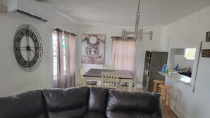 En stue med en sort lædersofa, et spiseområde med et træbord og stole, et køkken med en bar og et vægur.