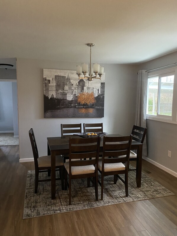 Un coin repas avec une table dressée pour quatre personnes, entourée de chaises en bois dotées d’assises rembourrées. Un lustre est suspendu au-dessus de la table, et un grand tableau représentant un paysage urbain est accroché au mur.