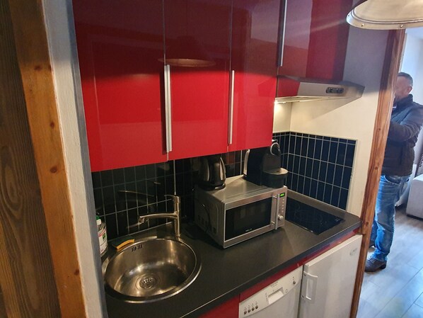 Eine kompakte Küche mit roten Schränken, einem Edelstahlspülbecken, einer Mikrowelle und einer Kaffeemaschine.