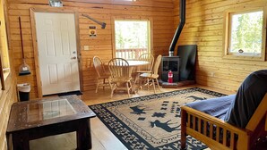 A cozy cabin interior with a wooden floor, a dining table with chairs, a bench, a sofa, a fireplace, and a window. - Fireweed Mountain Lodge - Bonzana Cabin (McCarthy)