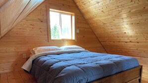 A wooden cabin bedroom with a bed, a window, and a sloped ceiling. - Fireweed Mountain Lodge - Bonzana Cabin (McCarthy)
