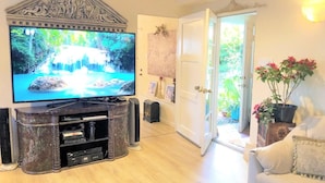 Uma sala de estar com uma televisão de tela plana grande sobre um suporte de madeira, uma porta branca que dá para uma área externa e um sofá branco com um vaso de planta.