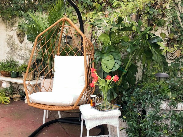 A wicker chair with a white cushion, a small table with a white cloth, and a vase with red flowers.