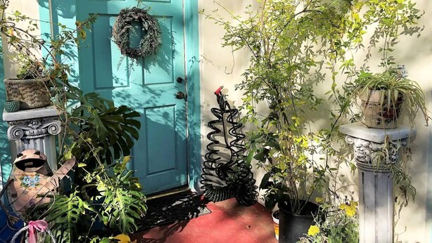 Uma porta turquesa adornada com uma grinalda, rodeada por diversos vasos de plantas e uma espiral metálica decorativa.