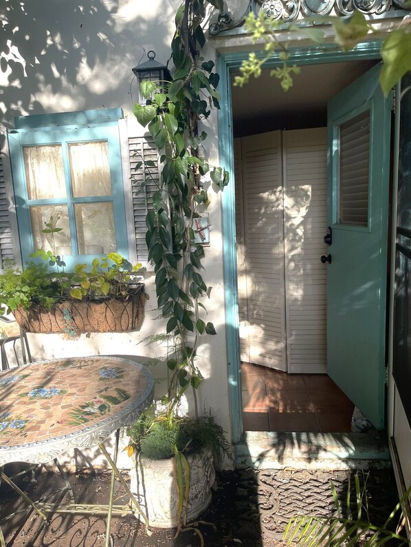 Una esquina de una casa con una puerta azul, una ventana con cortinas, una planta en maceta y una zona de patio con baldosas.