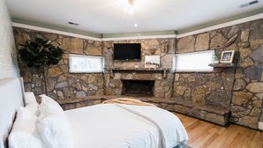 A bedroom with a stone fireplace, a bed with white bedding, and a television mounted above the fireplace.