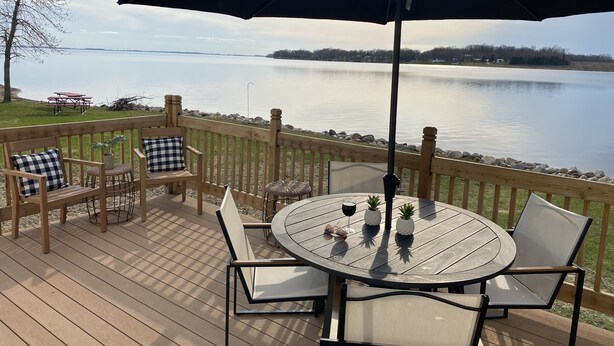 A wooden deck with a round table and chairs, an umbrella, and a view of a lake. - Enjoy Beautiful Sunsets on Lake Reno (Alexandria)