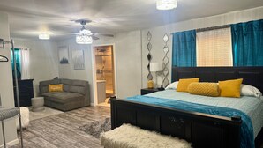 A bedroom with a bed, a bench, a sofa, a ceiling fan, and a nightstand with a lamp. - Upscale Abode  (Wichita)