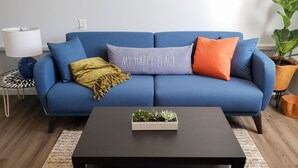 A blue sofa with a 'MY HAPPY PLACE' cushion, a yellow throw blanket, and a small plant on a black coffee table. - Cozy, modern 1-bedroom guest suite with backyard ocean view! (Pacifica)