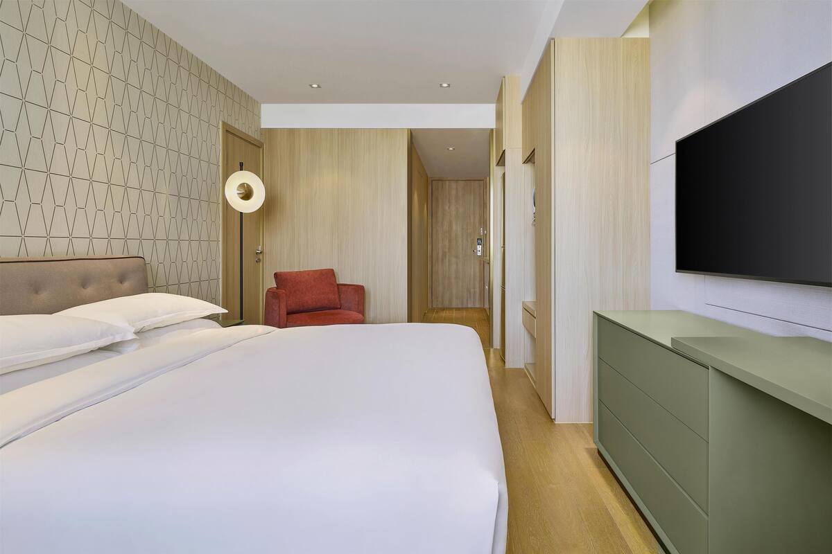 A modern hotel room with a large bed, a red chair, a TV mounted on the wall, and a wooden door.