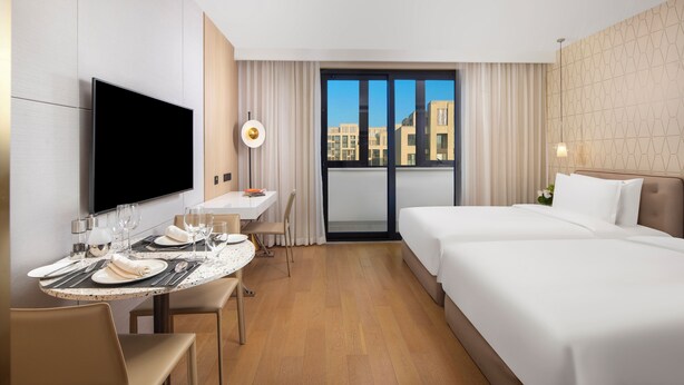 A hotel room with a bed, a desk with a chair, a television, and a large window with a view of buildings. - Hyatt Place Shanghai Lingang (Shanghai)