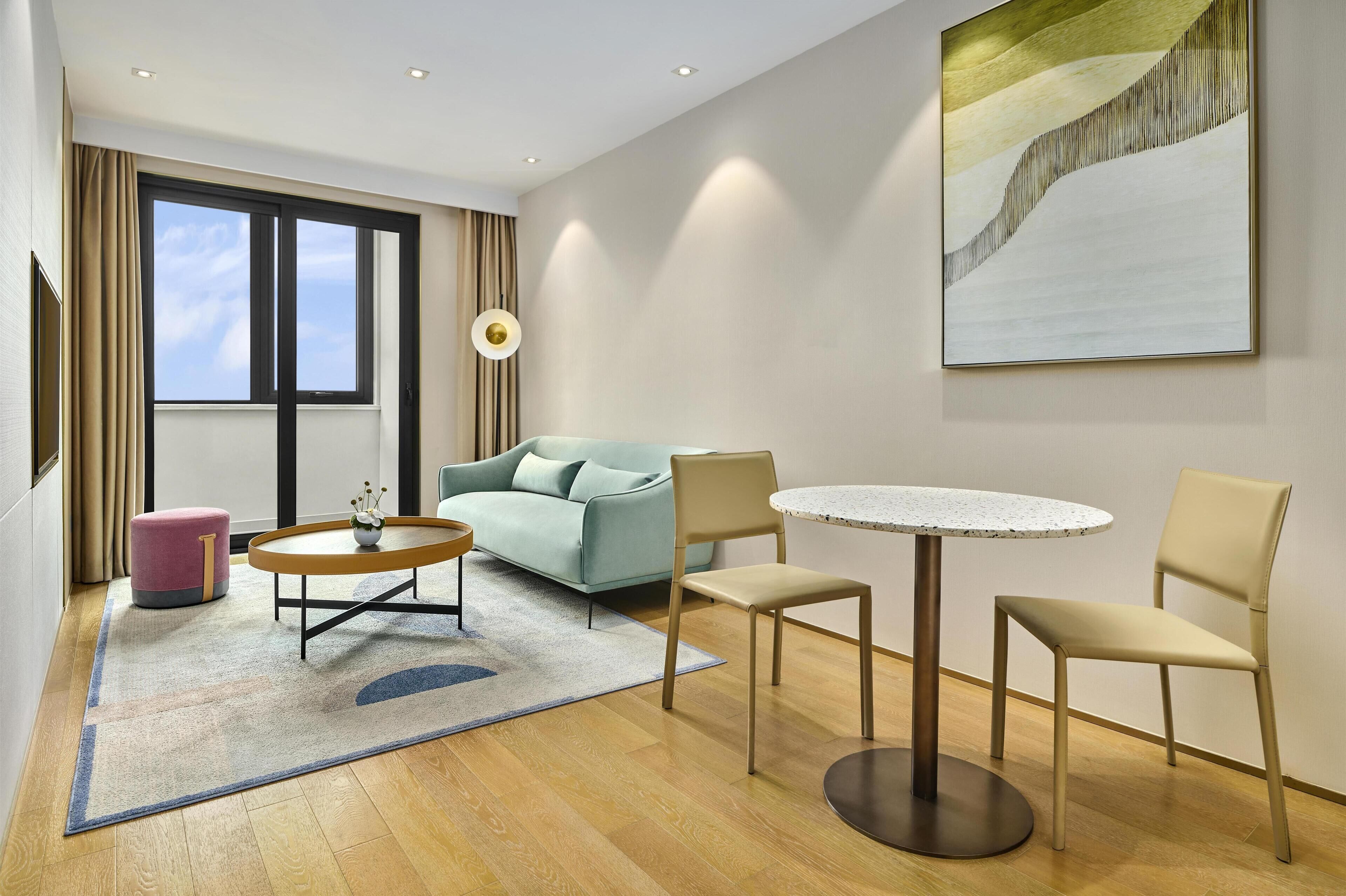 A modern hotel room with a sofa, round table, chairs, and a large abstract painting.