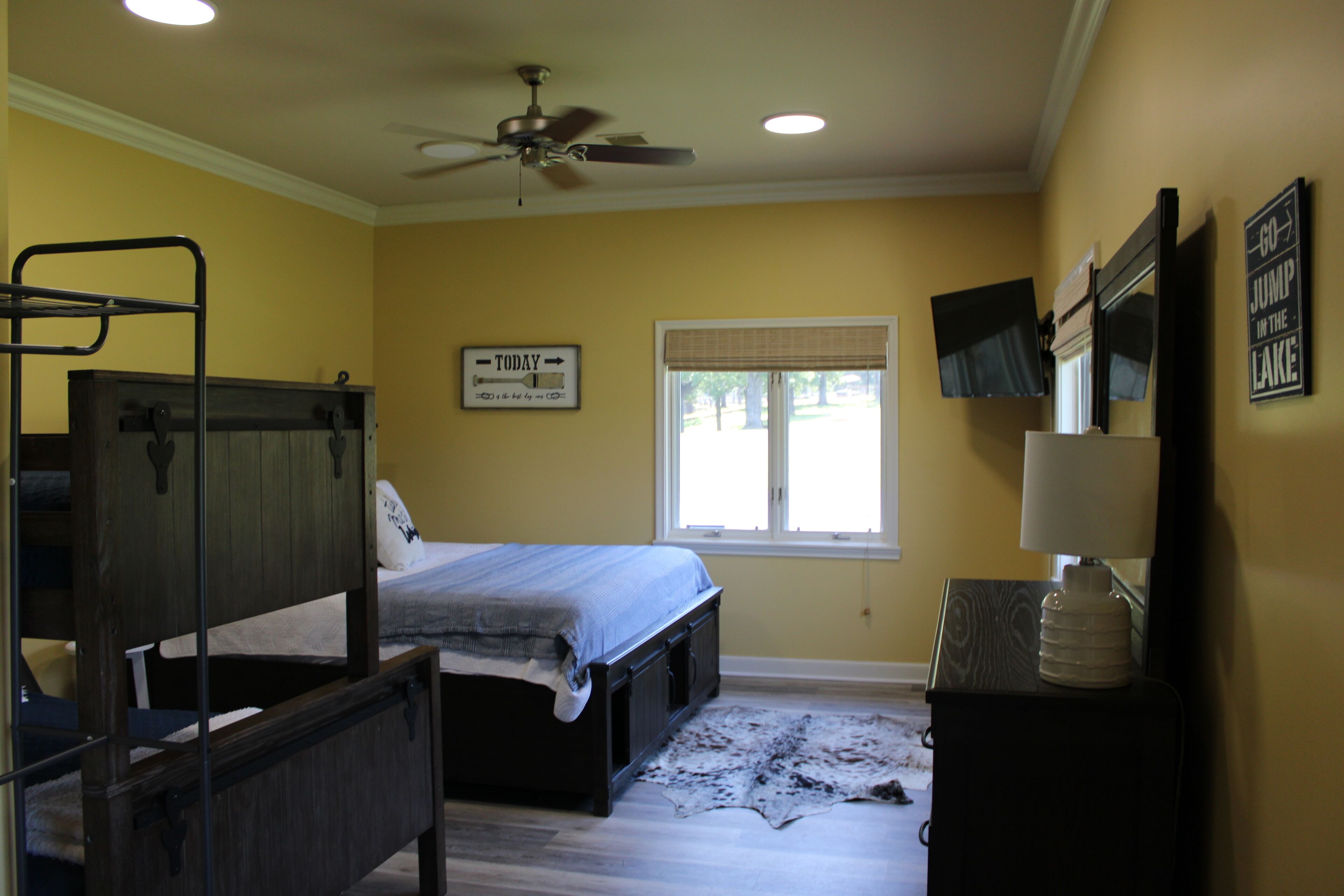 A bedroom with a bunk bed, a single bed, a ceiling fan, a window with blinds, a television, and a water dispenser.