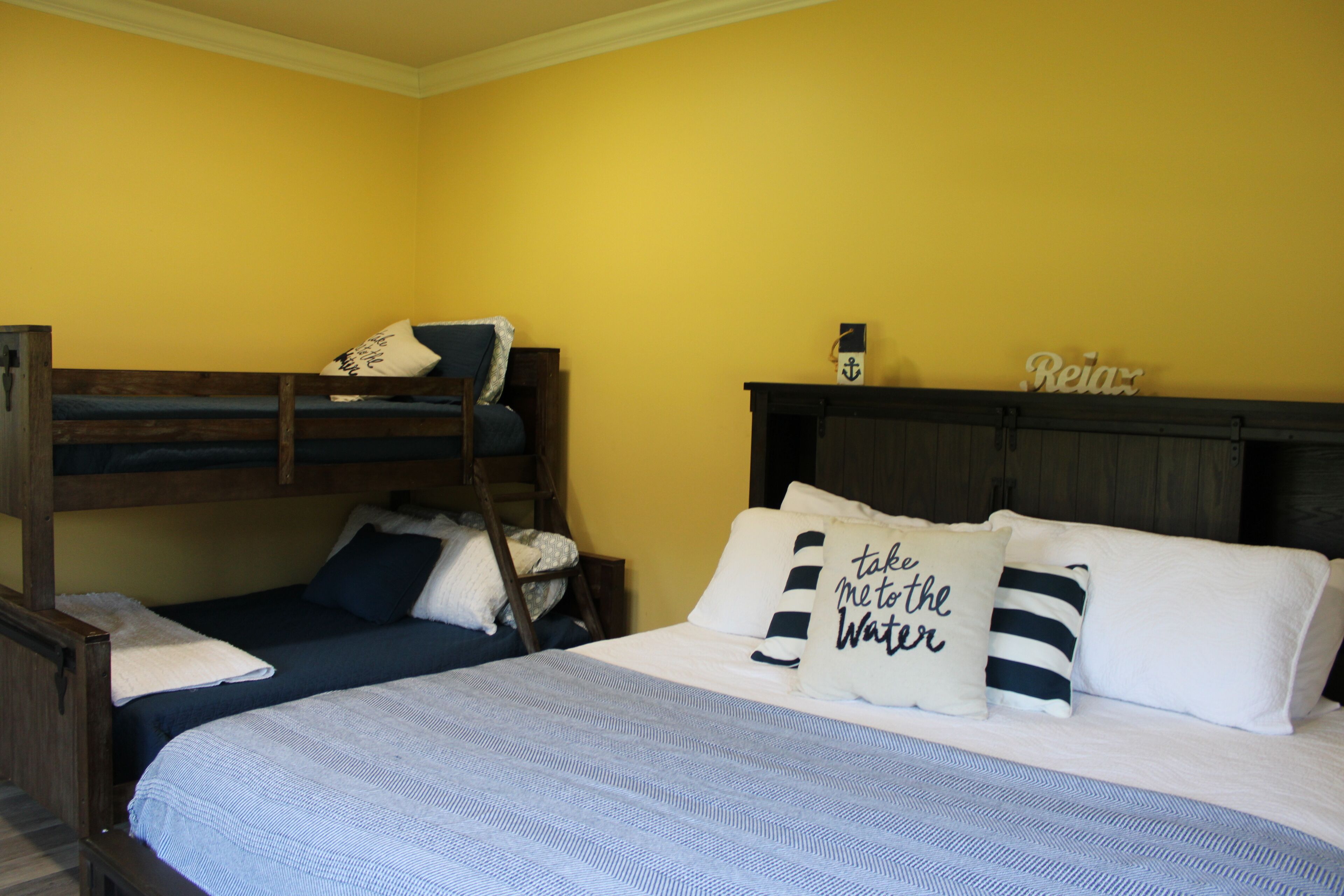 Une chambre avec un lit superposé, un lit simple avec une literie à rayures et un mur sur lequel est accroché un panneau portant l’inscription « Reia ».