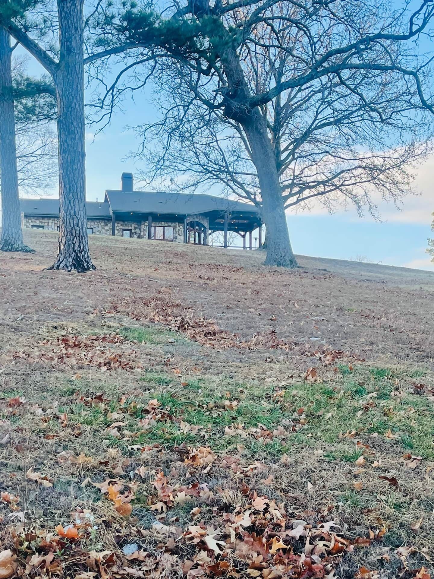 A building with a stone facade and a covered outdoor area, surrounded by trees and a grassy field with scattered leaves.