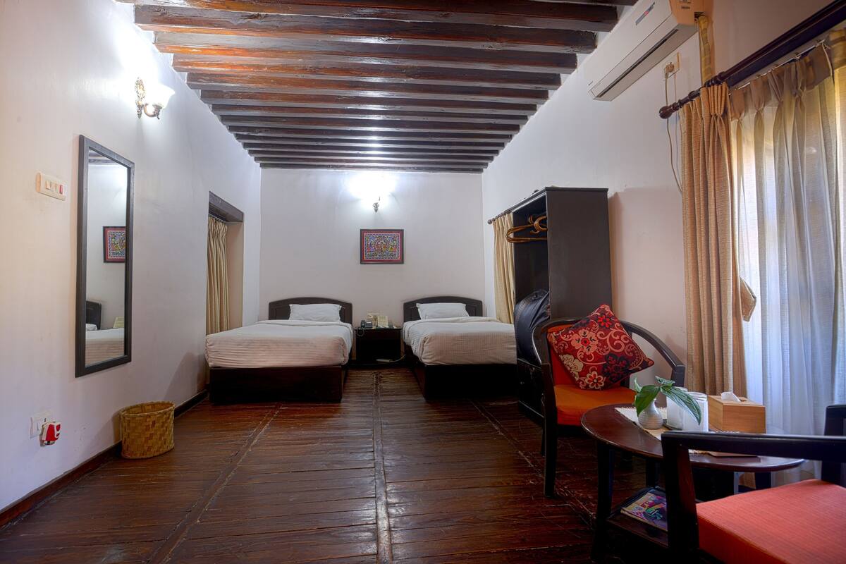 A room with two beds, a wooden ceiling, a mirror, a small table with a plant, and a chair.
