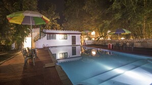 A swimming pool with a small building and outdoor seating area, surrounded by trees and umbrellas. - Krishna Jungle Resort  (Bichhiya)
