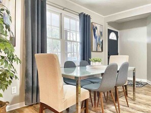 Un comedor con una mesa de vidrio, cuatro sillas, una planta en maceta y una ventana con cortinas.