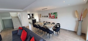 A modern living room with a glass dining table, black chairs, and a grey sofa with red cushions. A staircase leads to another room.