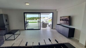 A modern living room with a grey sofa, a coffee table, a TV on a cabinet, and a view of a pool and greenery outside. - Sky Villa by Laranjal Rentals (Albufeira)