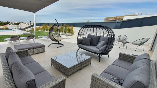 An outdoor patio with wicker furniture, a hanging chair, and a view of a swimming pool and buildings in the background. - Sky Villa by Laranjal Rentals (Albufeira)