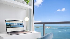 A laptop on a white desk with a view of a coastal landscape through a balcony.