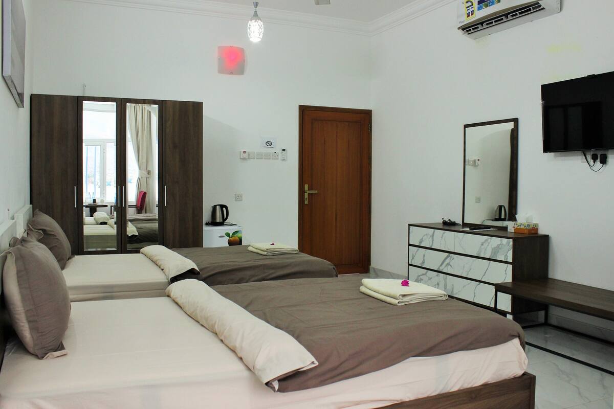 A hotel room with two beds, a wooden wardrobe, a marble-tiled dresser, a flat-screen TV, and a door with a handle.