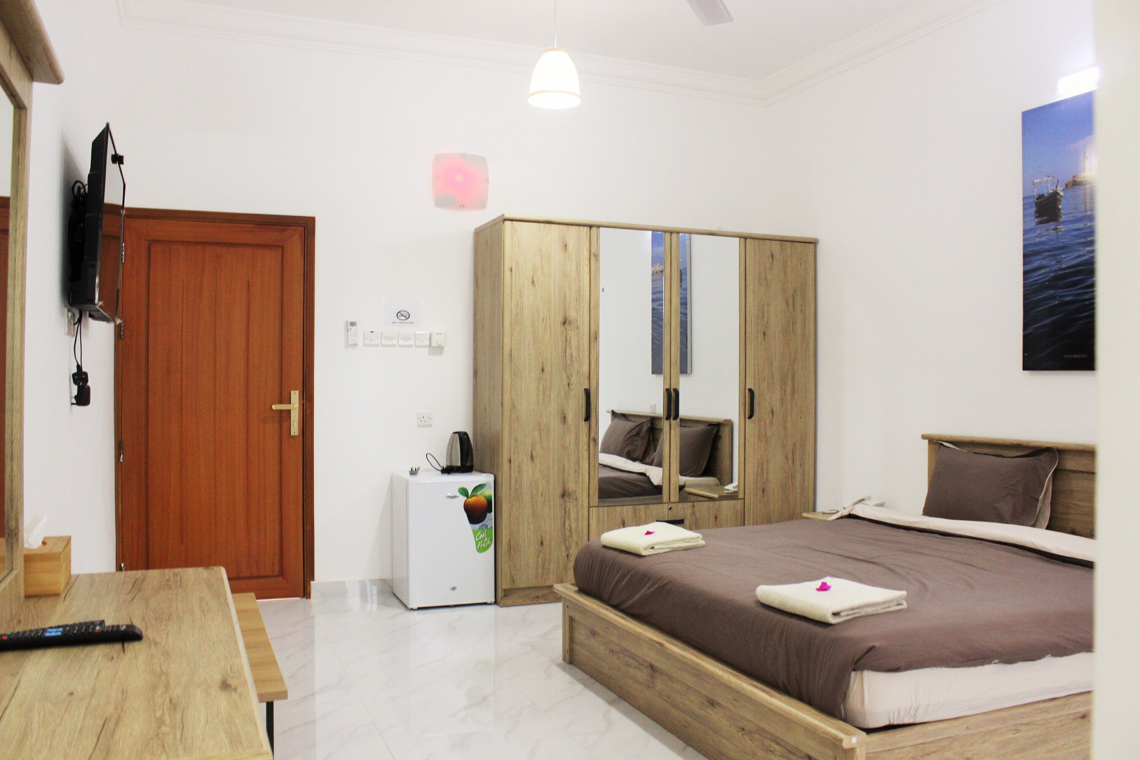 Une chambre avec une armoire en bois, un lit recouvert d’une couverture marron, un petit réfrigérateur blanc et un téléviseur fixé au mur.