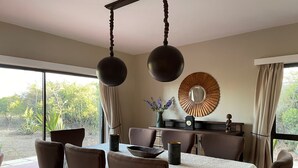 Un comedor con una mesa preparada para cuatro personas, rodeada de sillas, una lámpara colgante, un espejo decorativo y vistas al exterior.