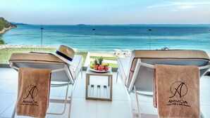 A balcony with two lounge chairs, each with a towel bearing the logo "AMMOA." A small table with fruits and a hat is between the chairs. The view includes a beach and the sea. - Ammoa Luxury Hotel & Spa Resort (Sithonia)