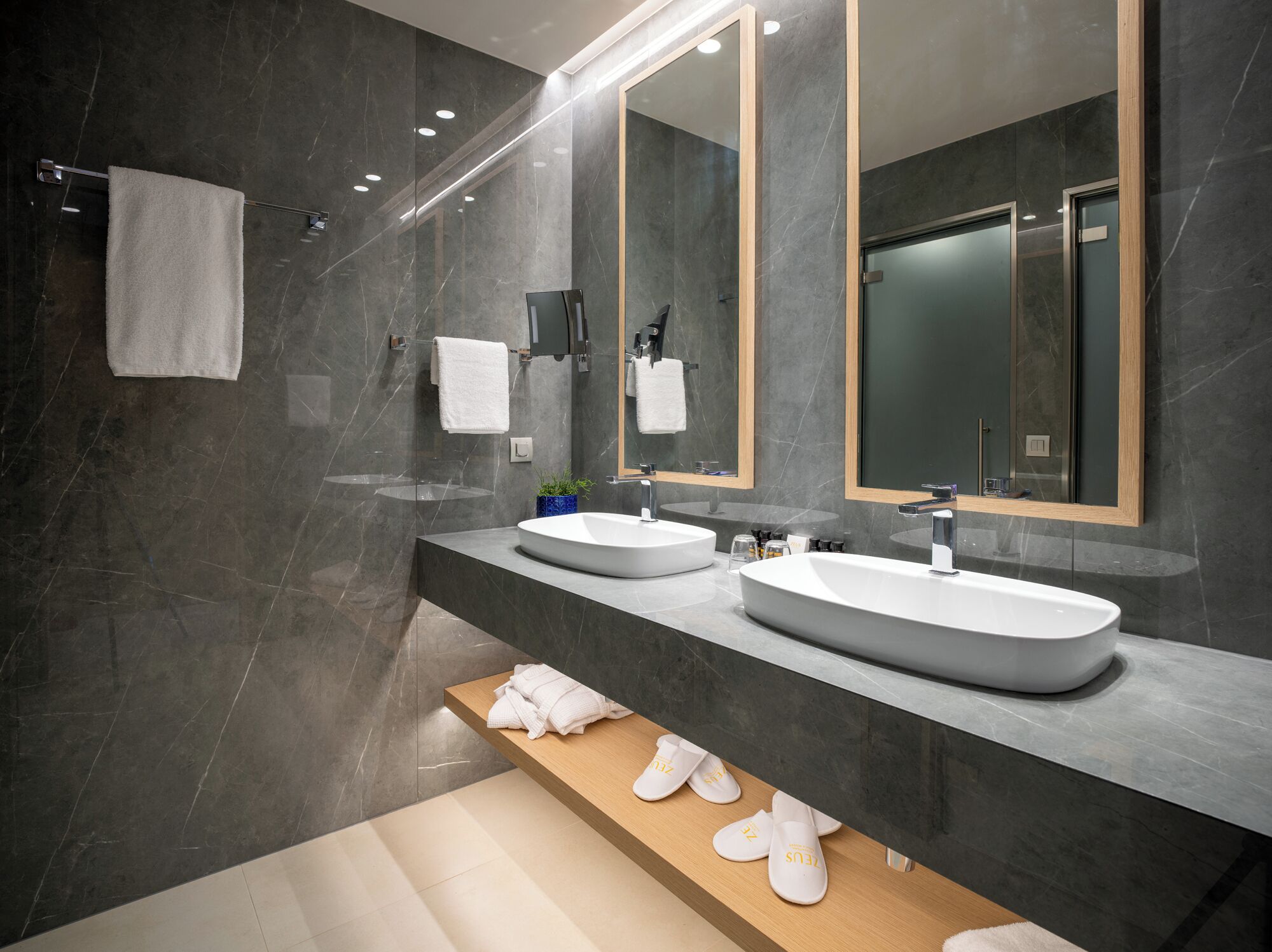 Un baño moderno con dos lavamanos, un espejo grande y un estante de madera con toallas y pantuflas.