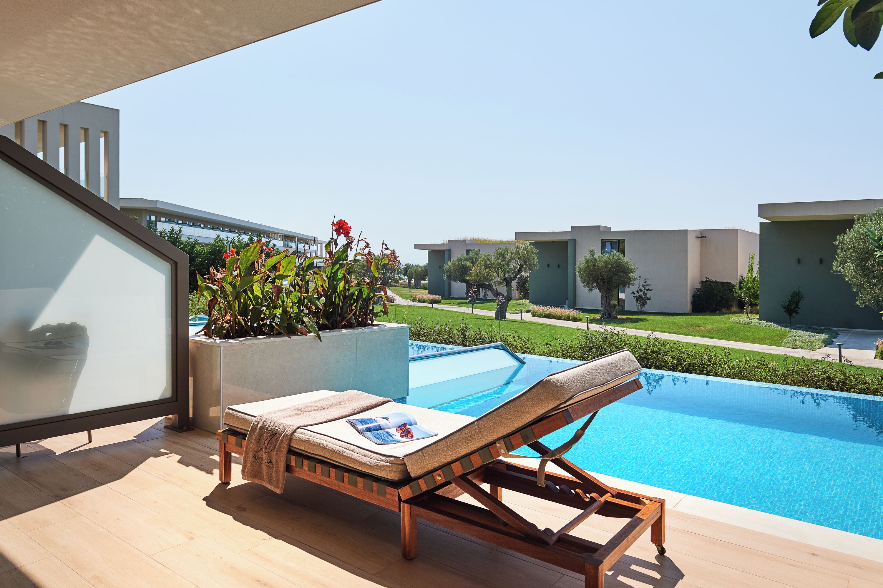 A modern outdoor pool area with a lounge chair and a magazine, surrounded by a well-maintained garden and a clear view of the sky.
