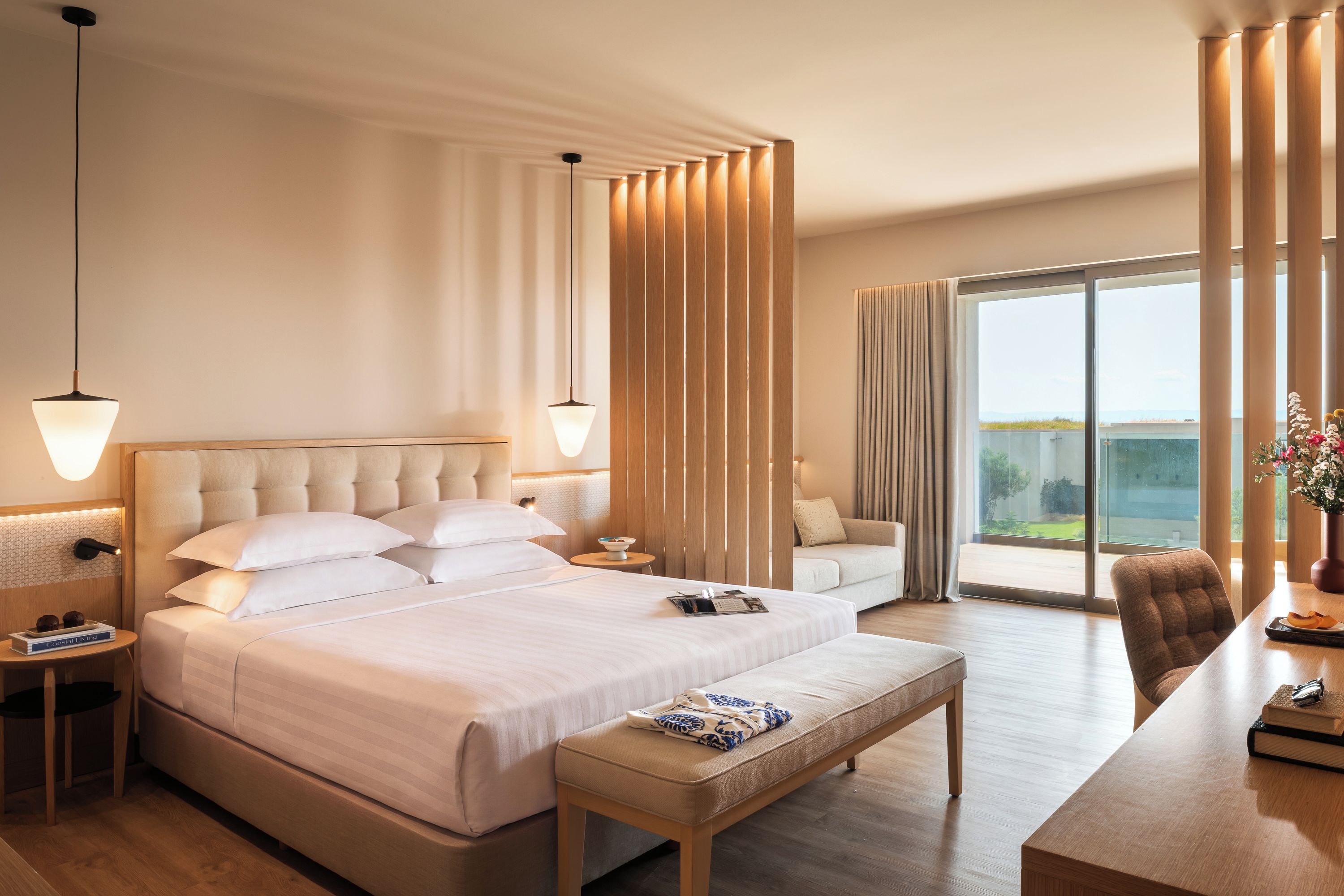 Quarto de hotel com uma cama grande, área de estar com sofá e poltronas, mesa de jantar e vista para o exterior.