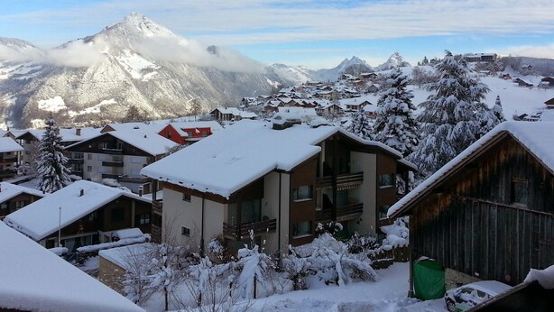 Un pueblo cubierto de nieve en la montaña, con edificios de estilo chalet y un pico destacado al fondo, bajo un cielo despejado.