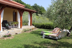 A covered outdoor seating area with lounge chairs and a table, surrounded by greenery and a stone patio.