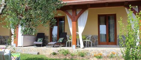 A covered outdoor seating area with a stone floor, surrounded by greenery and a small garden.