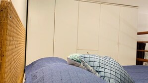 Uma cama com roupa de cama azul e cabeceira de tecido, um guarda-roupa com puxador e uma prateleira de madeira.