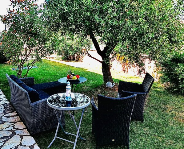 An outdoor seating area with wicker furniture and a round table, surrounded by a well-maintained lawn and trees.