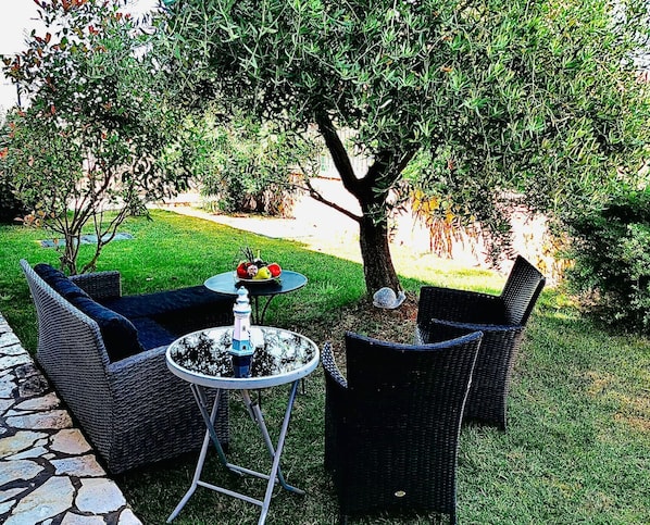 An outdoor seating area with wicker furniture and a round table, surrounded by a well-maintained lawn and trees.