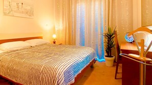 A hotel room with a bed, a nightstand with a lamp, a potted plant, a desk with a chair, and a window with curtains. - Vip Luxurious Apartment (Medulin)