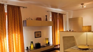 A modern living room with a sofa, a dining table, and a TV. There are curtains, a lamp, and a shelf with decorative items.