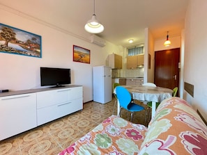 Un espace de vie compact comprenant un coin cuisine, un espace repas avec une table et des chaises, un canapé, un téléviseur posé sur un meuble et des œuvres d’art accrochées aux murs.