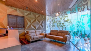 Un interior moderno con techo de madera, diseño geométrico en la pared, un sofá, un mostrador de recepción blanco y una pared de vidrio con un diseño decorativo.
