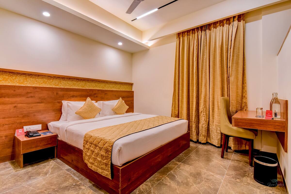 Une chambre d’hôtel avec une tête de lit en bois, un lit avec du linge de maison blanc, un bureau avec une chaise et une grande fenêtre avec des rideaux.