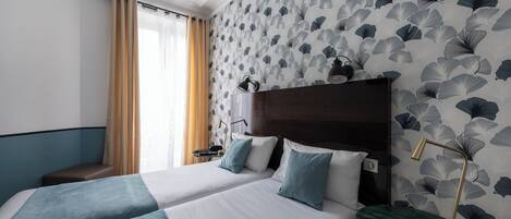 Habitación de hotel con dos camas, cabecero de madera, mesita de noche verde, escritorio y ventana con cortinas.