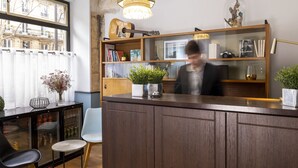 Un mostrador de recepción con un armario de madera, una vitrina de cristal con libros y objetos decorativos, una planta en maceta y vistas de un edificio a través de una ventana.