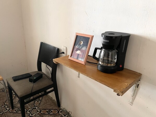A small wooden table with a black coffee maker and a framed picture. A black chair is positioned at the table.