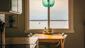 A dining area with a view of the sea through a large window, a table with chairs, and a hanging green light fixture. - Bayside Apartment #4- Top Floor Waterfront Views above Boulevard Park (Bellingham)