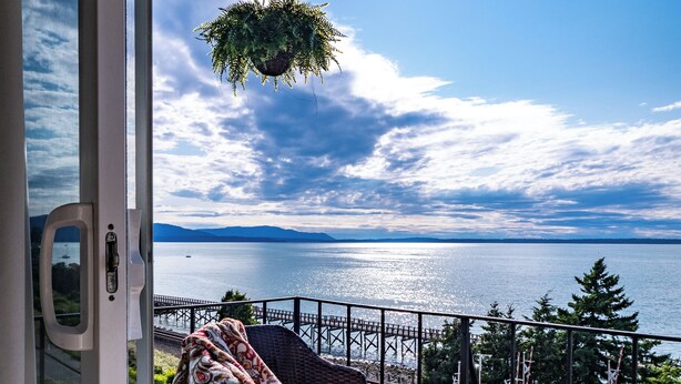 A balcony with wicker furniture, a hanging plant, and a view of the sea and mountains. - Bayside Apartment #4- Top Floor Waterfront Views above Boulevard Park (Bellingham)
